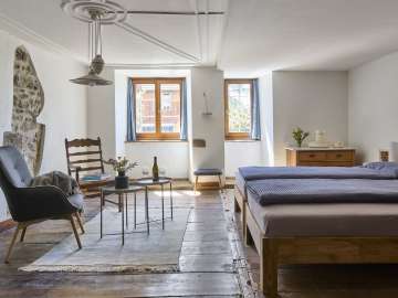 Dans une chambre au sol en bois, il y a un lit double, un fauteuil et une chaise en bois. Dans le mur crépi blanc, il y a un trou où le mur historique est mis à nu.