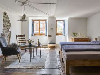 Baderhaus Neunkirch, vacances dans un monument historique Dans une chambre au sol en bois, il y a un lit double, un fauteuil et une chaise en bois. Dans le mur crépi blanc, il y a un trou où le mur historique est mis à nu.