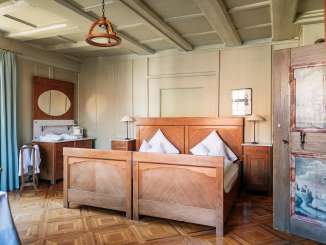 Chambre de l'auberge Hirschen Stammheim Un lit double en bois avec des draps blancs. Un vieux parquet en filigrane. Les murs sont beiges et la porte en bois a des peintures paysagères.