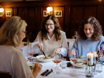 Drei Freundinnen sitzen an einem weiss gedeckten Tisch. Darauf stehen Weingläser mit Rosé und ein Fischmenü.