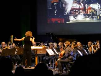 Une chef d'orchestre se tient sur scène avec l'orchestre. Derrière eux, on aperçoit une locomotive à vapeur sur un écran.