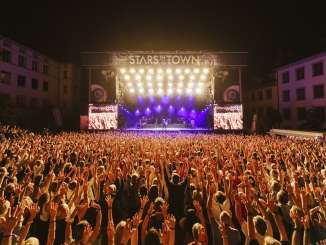 Des stars dans la ville De nombreuses personnes, les bras en l'air, assistent à un concert avec un groupe sur une grande scène.