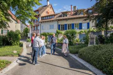 Visite guidée de Hallau Une femme en costume traditionnel se tient devant un groupe et raconte des histoires. Le groupe se trouve dans une ruelle avec des maisons aux façades colorées.