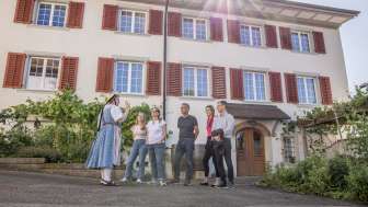 Visite guidée de Hallau Cinq personnes se tiennent devant une maison aux volets rouges. Un guide en costume traditionnel raconte des histoires de village.
