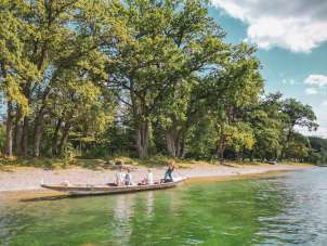 Promenade en weidling à Schaffhouse Trois personnes et un chien sont assis dans le weidling, un long bateau en bois. Une femme se tient à l'arrière et pique le long de la rive avec une plage de galets et des arbres.