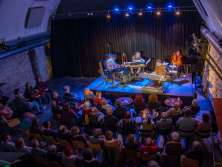 Une kleine Bühne avec quatre musiciens. La salle pleine écoute avec attention. L'éclairage est bleu et rouge.