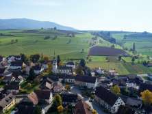 Vue sur le centre du village de Schleitheim avec une église au milieu. Derrière le village, il y a de légères collines avec des prairies et des champs.