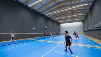 Une salle avec quatre terrains de badminton bleus. Des personnes y jouent au badminton.