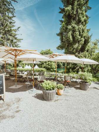 Des chaises et des tables en bois sont dressées et des parasols blancs offrent de l'ombre. La terrasse du restaurant est située directement au bord du Rhin et entourée de verdure.