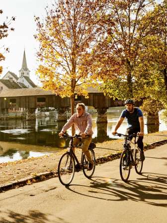 Faire du vélo à Diessenhofen Deux amis se promènent à vélo le long du Rhin. Sur la rive, il y a des arbres dorés et un pont en bois qui enjambe le Rhin.