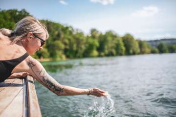 Une femme porte un maillot de bain noir et des lunettes de soleil. Elle est allongée sur un bateau en bois et tient une main dans l'eau.