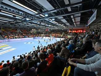 Un terrain de handball avec des gradins remplis de spectateurs. Le terrain est bleu. Sur une bannière, on peut lire l'inscription "BBC Arena".
