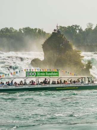 Un bateau vert rempli de personnes. Le panneau sur le toit du bateau indique : 30 min de croisière. Le bateau porte également l'inscription www.schiffmaendli.ch. En arrière-plan, on peut voir un rocher.