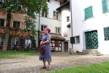 Une femme est vêtue d'un tablier et d'une robe bleu violacé et porte un foulard pourpre sur la tête. Elle se tient dans une cour intérieure.
