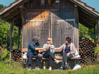 Trois personnes sont assises sur un banc en bois devant un Räbhüüsli dans les vignes. Ils trinquent avec un verre de vin blanc.