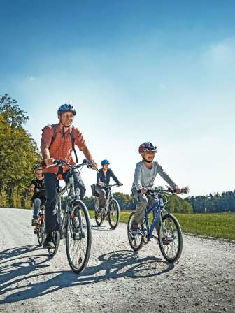 Tour des bords Une famille roule à vélo sur un chemin de terre. Autour d'eux, il y a des prés, des champs et une forêt.