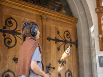 Schallhausen Musiktrail Une femme porte des écouteurs noirs "over the ears". Elle se tient devant la porte en bois d'une église. Elle porte un pull en laine marron sur l'épaule.