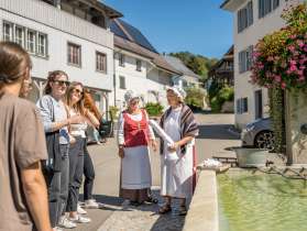 Près d'une fontaine du village, deux femmes sont déguisées en « Wöschwiiber », vêtues d'une robe et d'une coiffe. Un groupe les écoute raconter des histoires.