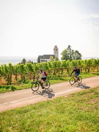 Faire du vélo à Hallau Une conductrice d'e-bike et un gravel-biker roulent ensemble sur une route à travers les vignes. On voit une église à l'arrière-plan.