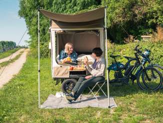 Dans un pré à côté de vignes, il y a un vélo-cargo avec une tente dépliée. Une personne est couchée sur le lit et assise sur une chaise de camping devant.