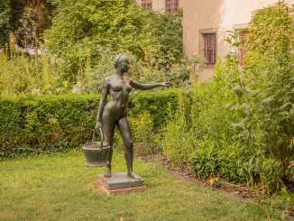 Une statue de femme portant un panier dans un parc. A l'arrière-plan, on voit la façade d'une maison brune.