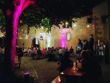 Bancs de fête dans une cour intérieure illuminée. Les bancs sont remplis de gens. Une kleine Bühne installée. Un musicien y joue de la musique.