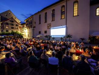 De nombreuses personnes sont assises sur une place devant une église et regardent un film qui sera projeté sur la façade de l'église,