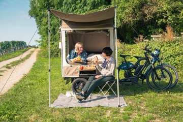 Dans un pré à côté de vignes, il y a un vélo-cargo avec une tente dépliée. Une personne est couchée sur le lit et assise sur une chaise de camping devant.