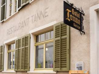 Sur la façade d'une maison, on peut lire en gros "Restaurant Tanne". Il a des volets verts et une enseigne noire en fer avec des lettres dorées "Hotel Tanne".