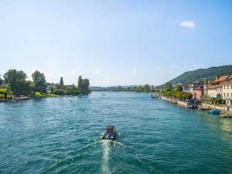 Un bateau et plusieurs stand up paddlers naviguent sur le Rhin. Sur la rive droite, on aperçoit le débarcadère de Stein am Rhein.