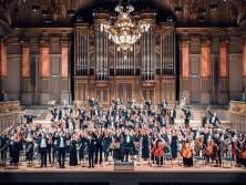 Auf einer Bühne vor einer prächtigen Orgel steht ein Orchester. Alle tragen etwas schwarzes.