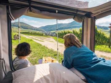 Une personne est allongée dans une tente installée sur un vélo cargo et contemple le paysage viticole. Une autre personne est assise devant la tente et prend son petit-déjeuner.