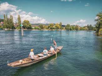 Trois personnes et un chien sont assis dans le weidling. Le bateau en bois descend tranquillement le Rhin. Une femme est debout, elle tient une rame et dirige le bateau.