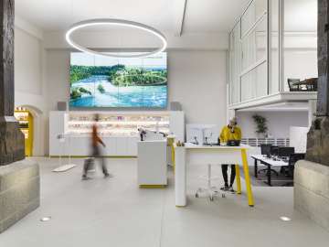 C'est la zone des guichets du Visitor Centre de Schaffhouse. Une femme en pull jaune moutarde est debout à une table haute et téléphone. Une autre personne se dirige vers le guichet libre.