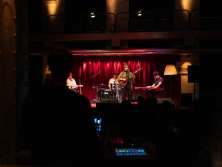 Un groupe de quatre personnes. La chanteuse principale est debout avec une guitare. La table de mixage est au premier plan, au centre de l'image.