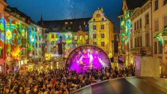 Des stars dans la ville Une place entre des maisons. Les façades sont éclairées en couleur. Au centre, une scène avec des musiciens/musiciennes. La place est remplie de gens.
