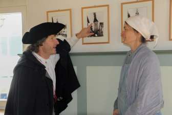 Une personne vêtue d'une cape noire et d'un chapeau noir montre une image avec une tour en feu. Une deuxième personne, vêtue d'une robe claire et d'un foulard blanc, écoute attentivement.