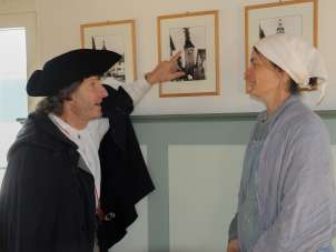 Une personne vêtue d'une cape noire et d'un chapeau noir montre une image avec une tour en feu. Une deuxième personne, vêtue d'une robe claire et d'un foulard blanc, écoute attentivement.