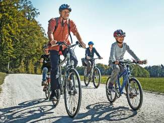 Une famille roule à vélo sur un chemin de terre. Autour d'eux, il y a des prés, des champs et une forêt.