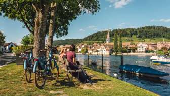 Faire du vélo à Stein am Rhein Deux amies sont assises sur un banc en bois au bord du Rhin avec vue sur Stein am Rhein. Derrière elles se trouvent deux vélos électriques.