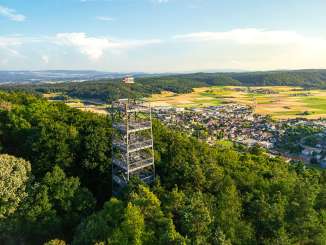 Randenturm de Béring Une tour métallique anguleuse émerge de la forêt sur une colline. En bas, on aperçoit le village Beringen.