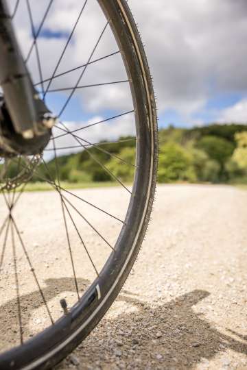 Partie d'une roue de vélo qui roule sur un chemin naturel. En arrière-plan, on voit la lisière de la forêt.