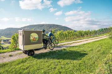 Deux amies se promènent dans les vignes avec leur vélo électrique. Sur un vélo-cargo, il y a une structure de tente dépliable.