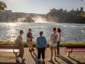 Un groupe de personnes se tient au bord du bassin des chutes du Rhin. Les chutes du Rhin sont visibles en arrière-plan. Tous écoutent attentivement une personne.