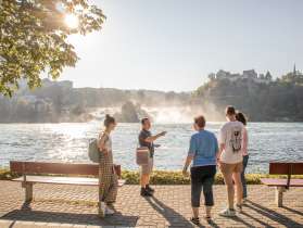 Un groupe de personnes se tient au bord du bassin des chutes du Rhin. Les chutes du Rhin sont visibles en arrière-plan. Tous écoutent attentivement une personne.