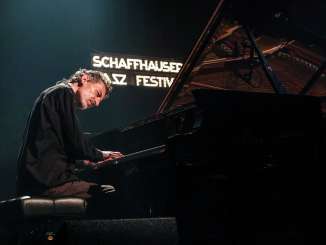 Festival de jazz de Schaffhouse Un homme est assis à un piano à queue et joue sur une scène. En arrière-plan, on voit un panneau lumineux avec l'inscription "Schaffhauser Jazz Festiaval".