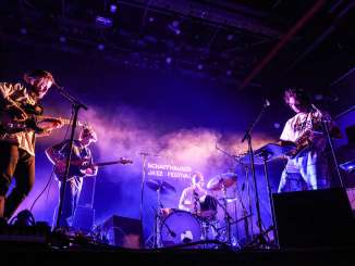 Festival de jazz de Schaffhouse Sur une scène, un batteur, deux guitaristes électriques et un saxophoniste sont éclairés par des projecteurs violets.