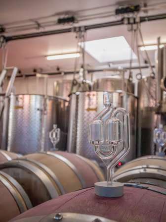 Une bonde de fermentation est plantée dans un fût en bois. Plusieurs cuves en acier se trouvent à l'arrière-plan.