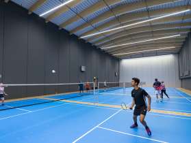 Une salle avec quatre terrains de badminton bleus. Des personnes y jouent au badminton.