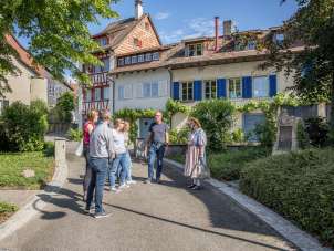 Une femme en costume traditionnel se tient devant un groupe et raconte des histoires. Le groupe se trouve dans une ruelle avec des maisons aux façades colorées.